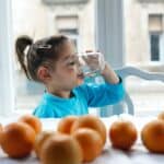 Child in blue shirt drinking water at windowsill with apples, promoting healthy hydration and nutrition.