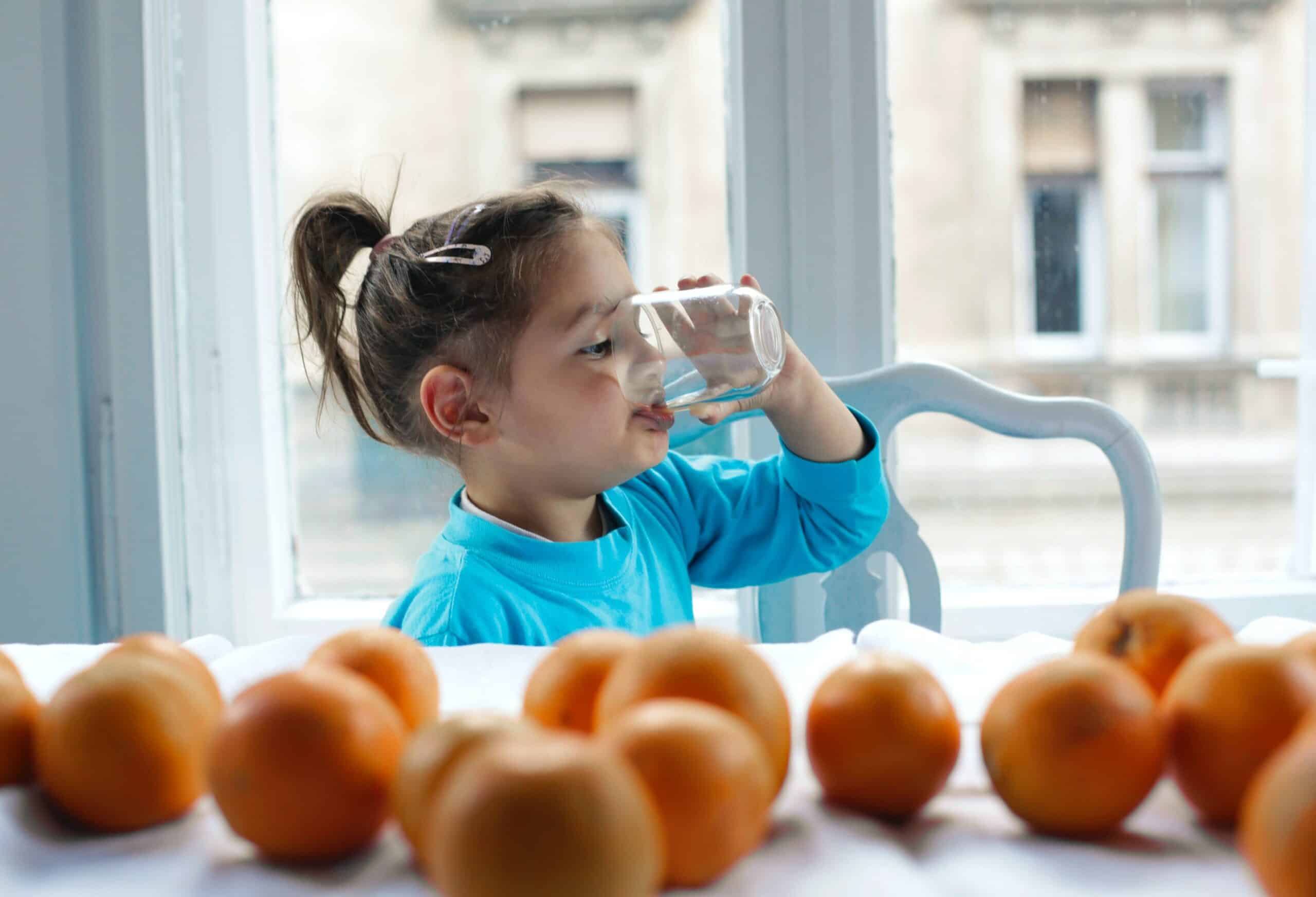 Child in blue shirt drinking water at windowsill with apples, promoting healthy hydration and nutrition.