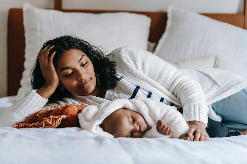 Mother and infant sleeping peacefully together on white bedding in bedroom.