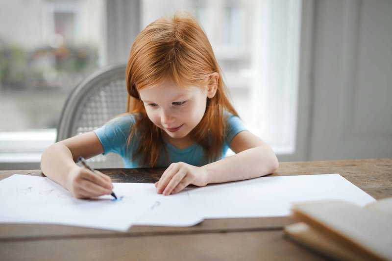 Young girl with red hair writing or drawing at desk near window in bright room.