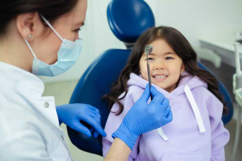 Pediatric dentist in mask and gloves examining smiling young girl in dental chair.