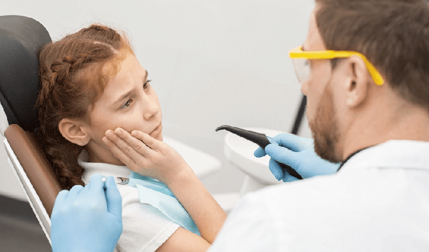 Young girl in dental chair touching her mouth while dentist examines her teeth with tools.