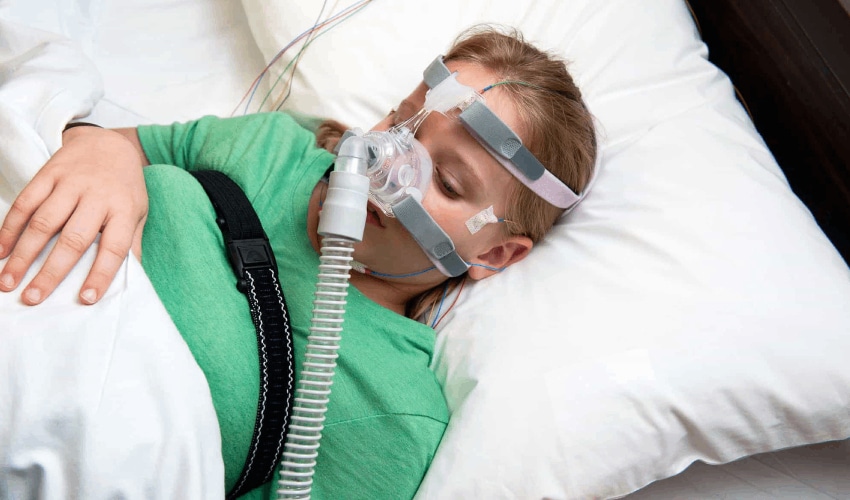 Child sleeping in bed wearing green pajamas and CPAP mask for sleep apnea treatment.