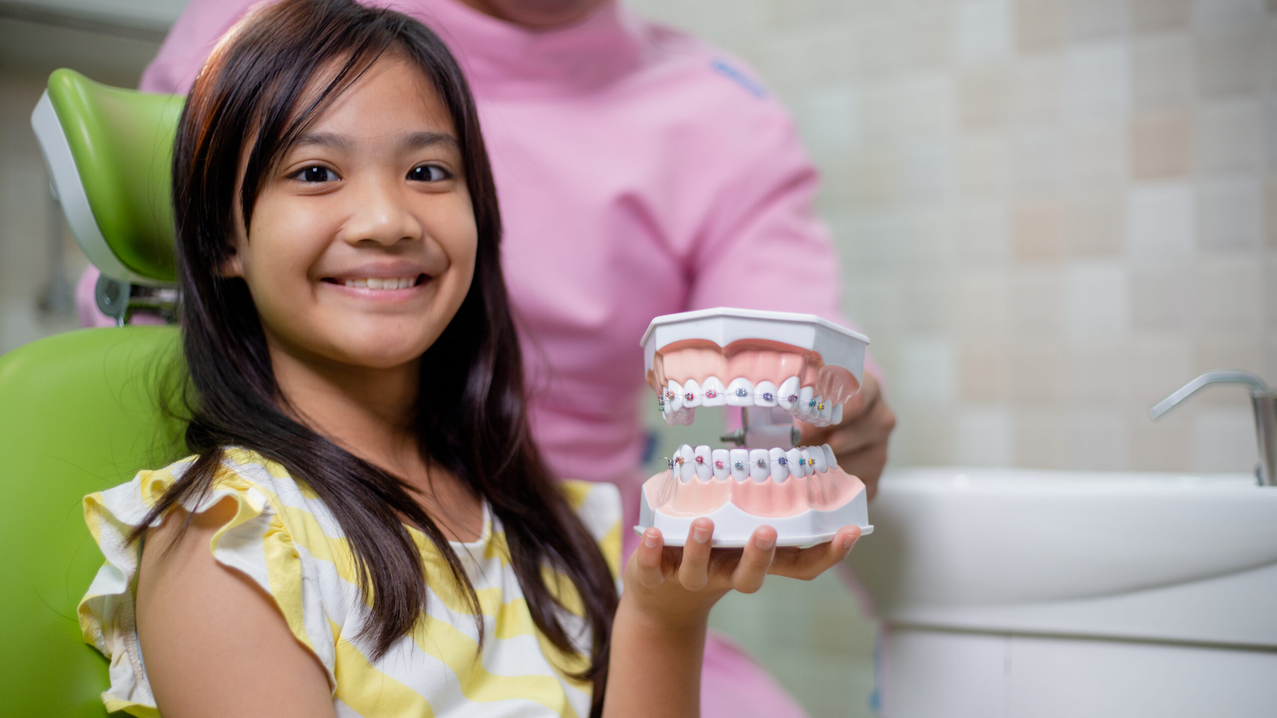 Young girl smiling in a dental chair while holding a model of teeth with braces during an orthodontic visit.