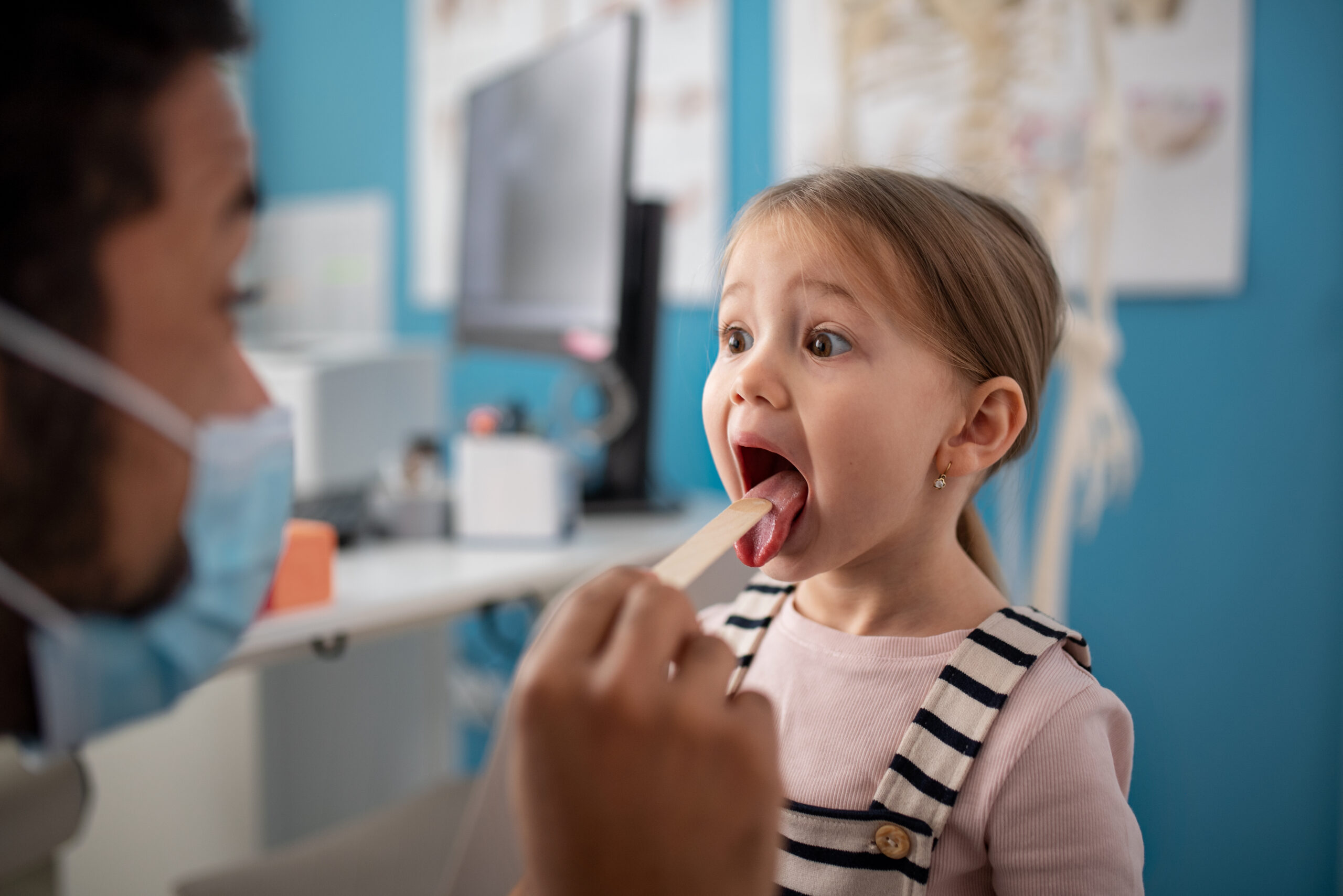 Young girl opening her mouth wide during a pediatric dental checkup with a masked provider using a tongue depressor
